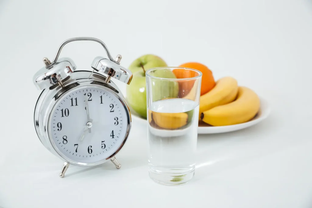 Alarm clock beside a glass of water and fresh fruits, symbolizing intermittent fasting for weight loss, meal timing, and healthy eating for beginners.