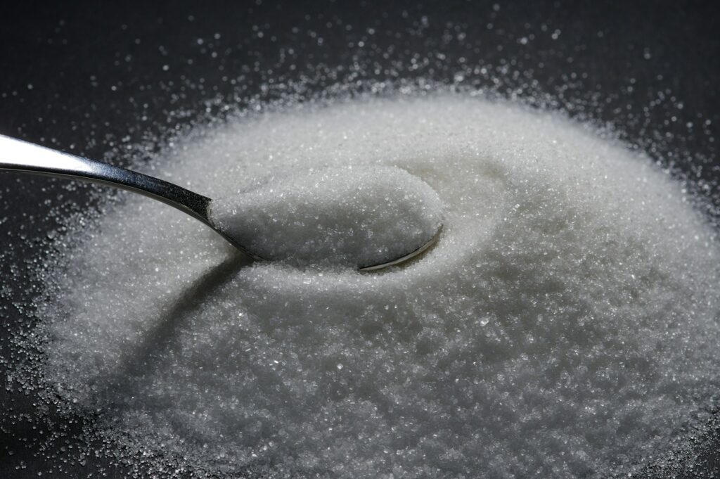 A metal spoon resting in a mound of white granulated sugar, with sugar crystals scattered on a dark surface in the background