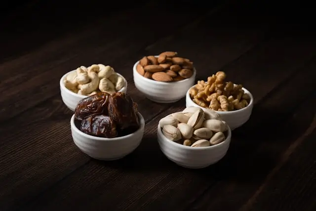 Small bowls of dates, pistachios, walnuts, almonds, and cashews on a wooden table, illustrating satisfying low-calorie snacks rich in protein, fiber, and healthy fats that help curb hunger.