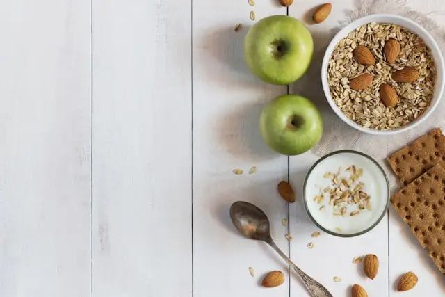 Healthy snack ingredients including green apples, oatmeal, yogurt, almonds, and whole grain crackers on a wooden table, illustrating nutritious low-calorie snacks that help control hunger between meals.