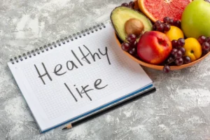 “Notebook with the words ‘Healthy Life’ beside a bowl of fresh fruits including avocado, apple, grapes, and grapefruit, symbolizing a healthy lifestyle, mental reset, and natural habits for improved focus and dopamine balance.
