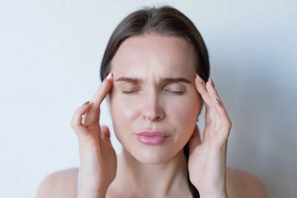 A woman with eyes closed and a pained expression, pressing her fingers to her temples as if experiencing a headache or dizziness. The image illustrates common detox symptoms to watch out for, showing the physical discomfort that can occur during a body cleanse.