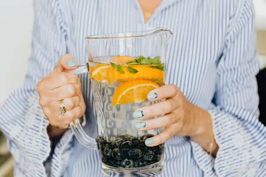 Woman holding a glass pitcher of infused water with orange slices, blueberries, and mint, representing natural detox habits for hormonal imbalance and what helps restore hormone balance naturally.